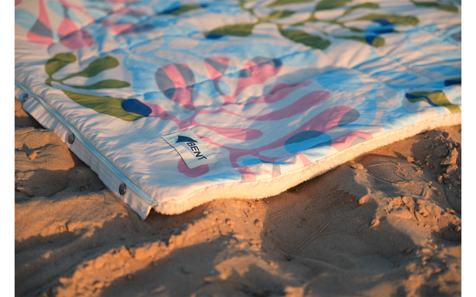 Coin d'un tissu coloré à motif de feuilles, bord cousu et œillets métalliques, posé sur du sable granuleux