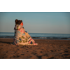 Personne aux cheveux relevés en chignon et portant un bandeau, enveloppée dans une couverture à motifs, assise sur une plage de sable avec la mer et un ciel dégagé en arrière-plan