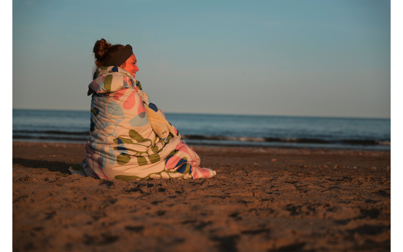 Personne aux cheveux relevés en chignon et portant un bandeau, enveloppée dans une couverture à motifs, assise sur une plage de sable avec la mer et un ciel dégagé en arrière-plan