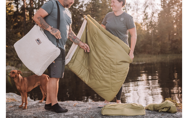 Deux personnes se tiennent sur un rocher au bord d’un lac en tenant une couverture verte; à gauche se trouve un chien brun; un tapis vert enroulé et un sac de couleur claire sont posés sur le rocher; en arrière-plan, arbres et eau