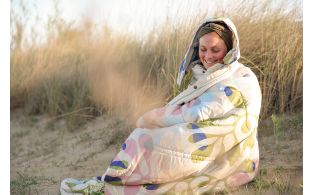 Personne enveloppée dans une couverture à motifs assise sur une dune de sable parmi des herbes de plage, sourit et regarde vers le bas