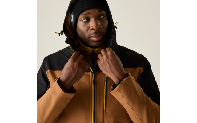 Personne portant une veste à capuche marron et noire et un bonnet noir, mains tenant la capuche, regard légèrement de côté, fond uni clair