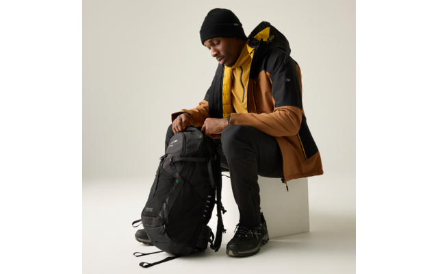 Personne assise sur un bloc blanc, porte un bonnet noir, une veste marron et noire avec doublure jaune, un pull jaune, un pantalon noir et des chaussures noires ; tient un sac à dos noir et regarde vers le bas