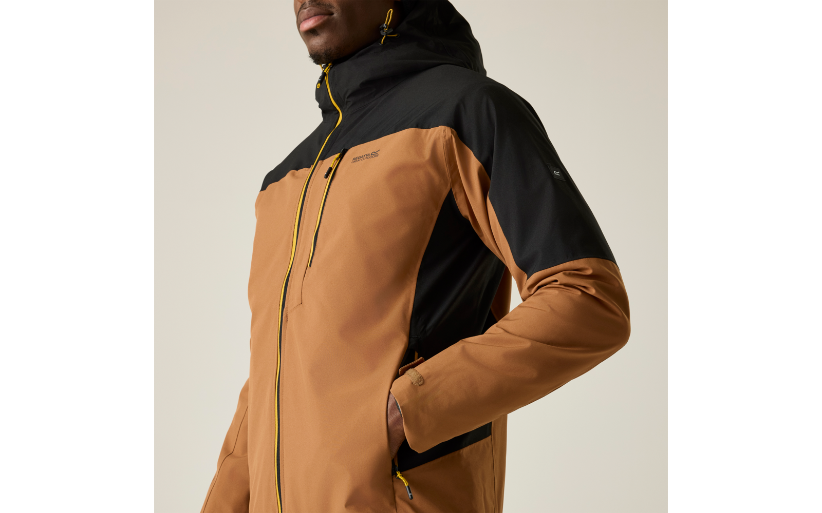 Gros plan sur le torse d'une personne portant une veste à capuche marron et noire avec fermetures éclair jaunes, main droite dans la poche, partie inférieure du visage visible