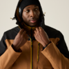 Personne portant une veste à capuche marron et noire et un bonnet noir, mains tenant la capuche, regard légèrement de côté, fond uni clair