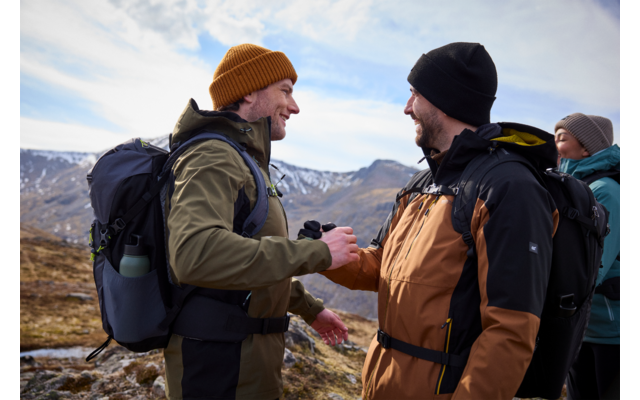 Zwei Männer mit Rucksäcken und Winterkleidung stehen draußen und sehen sich an, im Hintergrund Berge