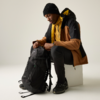 Personne assise sur un bloc blanc, porte un bonnet noir, une veste marron et noire avec doublure jaune, un pull jaune, un pantalon noir et des chaussures noires ; tient un sac à dos noir et regarde vers le bas