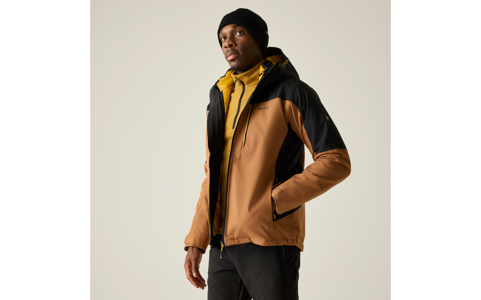Personne portant une veste d'extérieur bicolore (brun et noir) avec doublure jaune et un bonnet noir, mains dans les poches, regard légèrement vers le haut à droite, fond uni clair