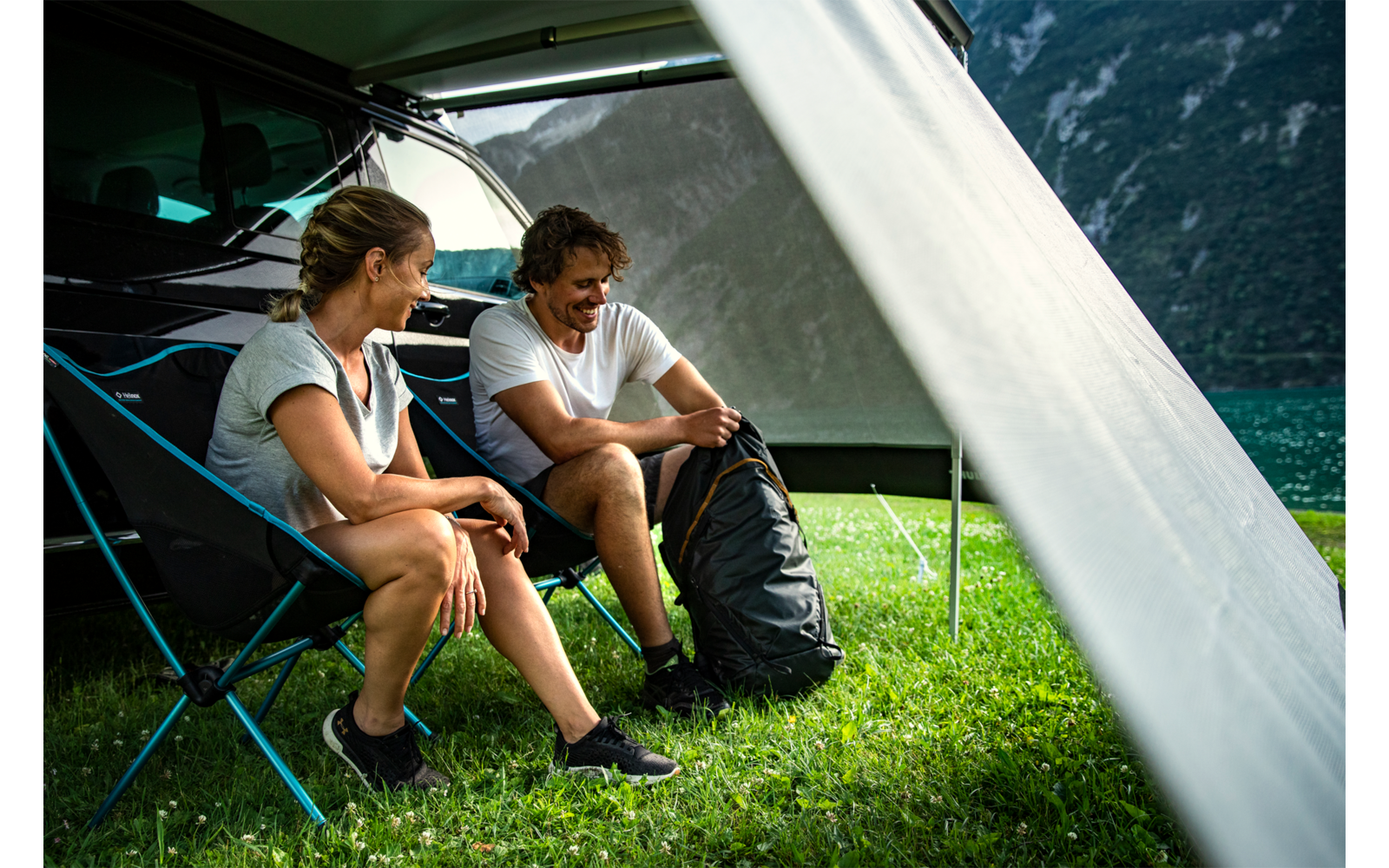 Zwei Personen sitzen auf Campingstühlen unter einer Markise neben einem Wohnmobil auf Gras; ein Rucksack neben ihnen; im Hintergrund See und Berge