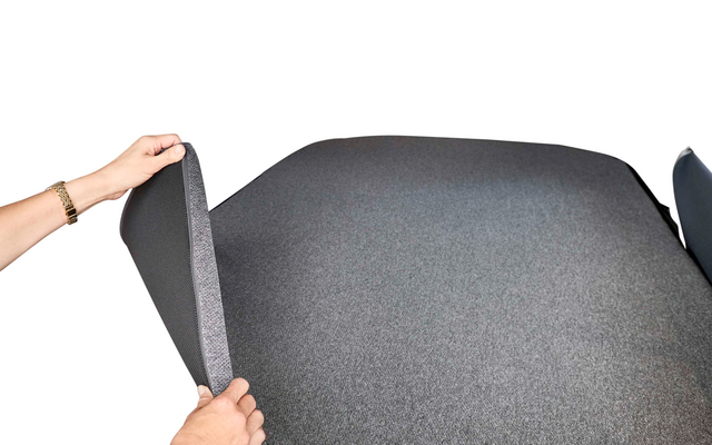 Zwei Hände heben einen dunkelgrauen Stoffbezug von einer dunkelgrauen gepolsterten Liegefläche; darunter ist eine hellgraue Trägerplatte mit geometrischem Muster zu sehen; helle textile Wände im Hintergrund