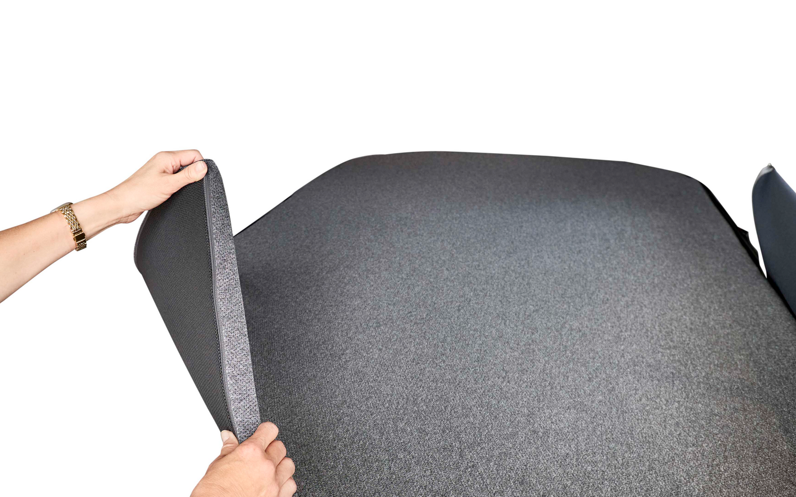 Zwei Hände heben einen dunkelgrauen Stoffbezug von einer dunkelgrauen gepolsterten Liegefläche; darunter ist eine hellgraue Trägerplatte mit geometrischem Muster zu sehen; helle textile Wände im Hintergrund