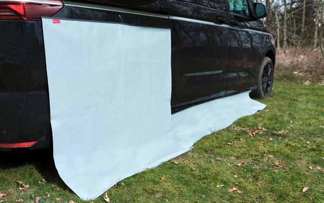 Dunkles Fahrzeug auf Gras, an der Seite hängt ein großes weißes Tuch, das auf dem Boden ausgebreitet ist; Bäume im Hintergrund