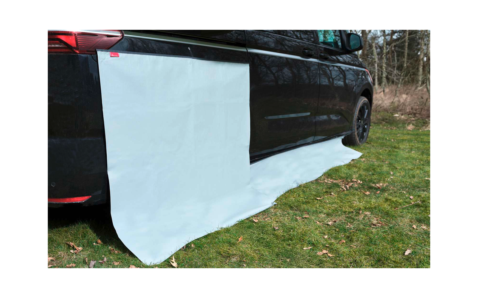 Dunkles Fahrzeug auf Gras, an der Seite hängt ein großes weißes Tuch, das auf dem Boden ausgebreitet ist; Bäume im Hintergrund