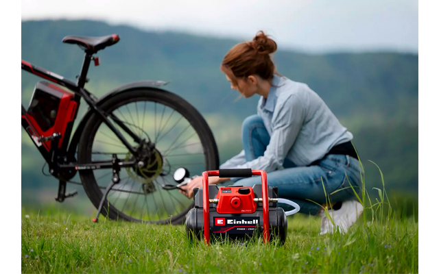 Frau kniet auf Wiese neben elektrischem Fahrrad und bedient roten tragbaren Kompressor; Berge im Hintergrund