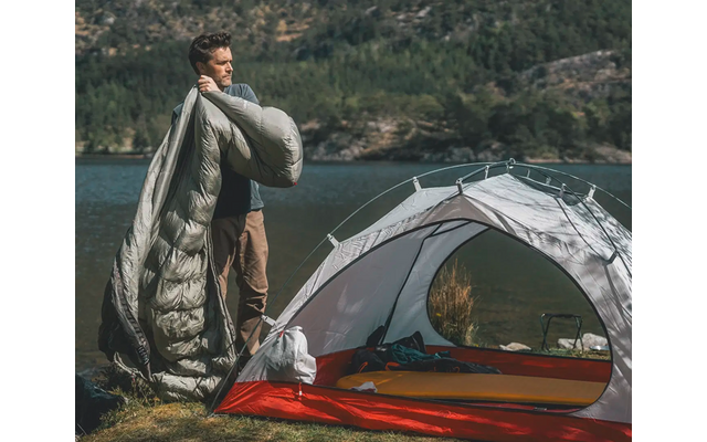 Mann steht neben offenem Zelt und hält Schlafsack in der Hand, im Hintergrund Berge und Wasser