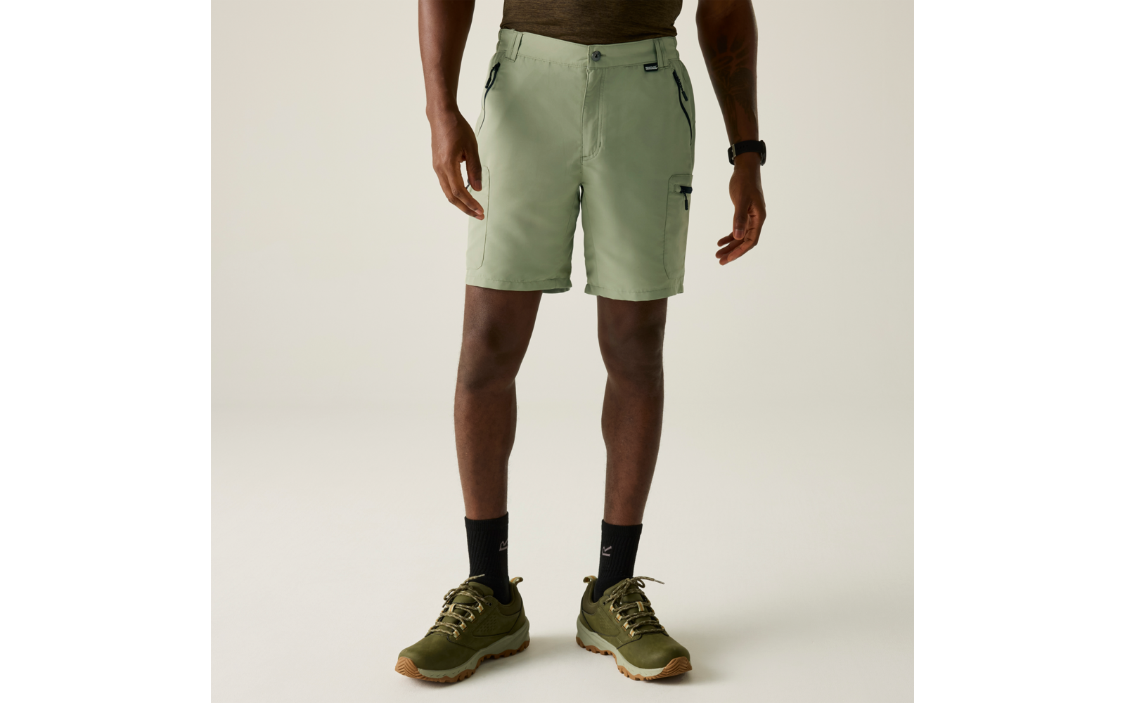 Person von der Hüfte bis zu den Füßen, trägt hellgrüne Shorts mit seitlichen Reißverschlusstaschen, schwarze Socken und olivgrüne Sportschuhe; Hände neben dem Körper