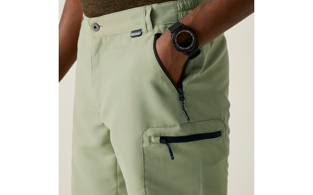 Nahaufnahme von Hüfte und Oberschenkel einer Person; Hand in Reißverschlusstasche einer hellgrünen Hose, zusätzliche Reißverschlusstasche am Oberschenkel, schwarze digitale Armbanduhr, braun-grünes Oberteil sichtbar