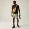 Homme debout sur fond clair, porte un T‑shirt vert foncé, short vert clair, sac beige en bandoulière, tient une gourde noire, chaussures de sport vertes