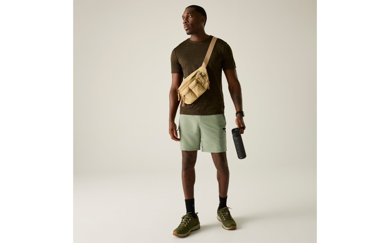 Homme debout sur fond clair, porte un T‑shirt vert foncé, short vert clair, sac beige en bandoulière, tient une gourde noire, chaussures de sport vertes