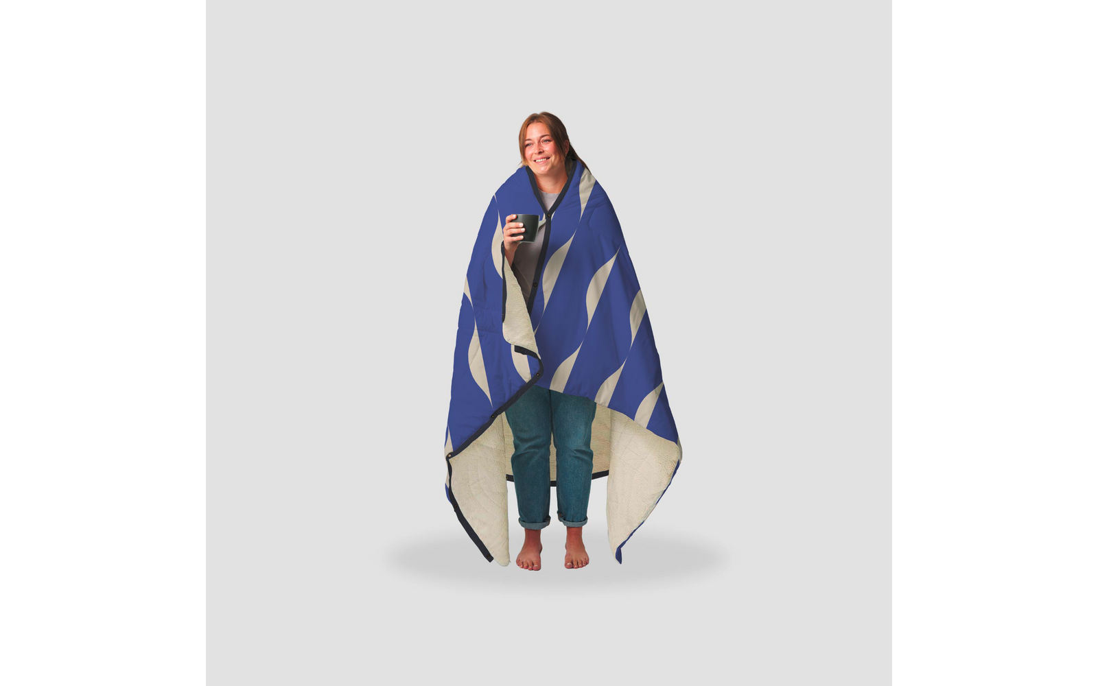 Person steht auf hellem Hintergrund, in eine blau-weiß gemusterte Decke gehüllt, hält eine Tasse, trägt hochgekrempelte Jeans und ist barfuß