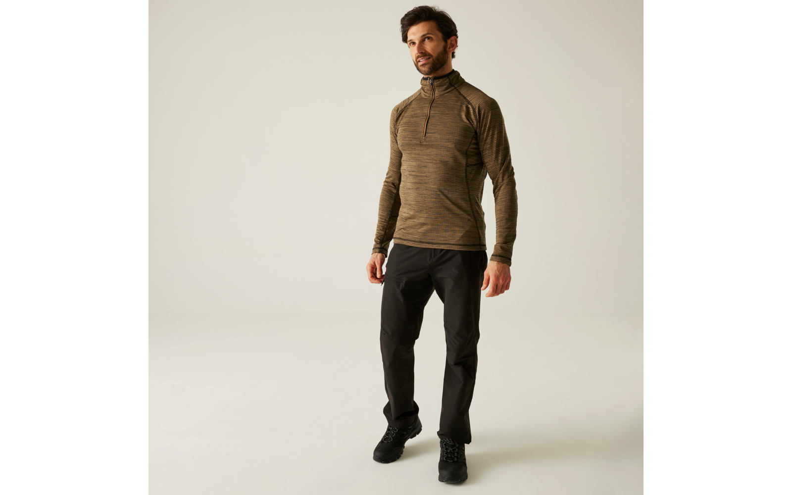 Ein Mann steht vor neutralem Hintergrund, trägt eine braune Jacke und schwarze Hose