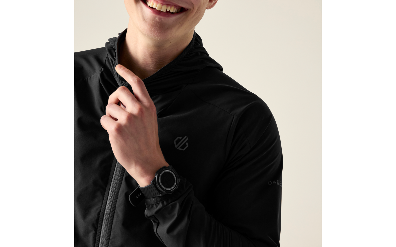 Nahaufnahme einer lächelnden Person, Oberkörper bis zum Kinn sichtbar, trägt schwarze Kapuzenjacke mit Reißverschluss, zeigt mit Zeigefinger auf die Brust, trägt schwarze Armbanduhr, heller Hintergrund