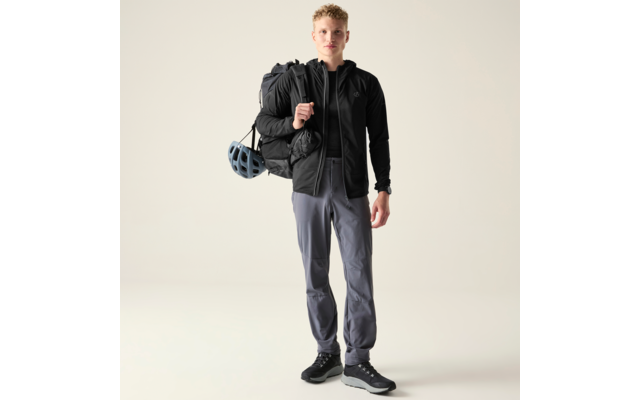 Person steht frontal vor hellem einfarbigem Hintergrund, trägt schwarze Kapuzenjacke mit Reißverschluss, graue Hose und dunkle Sneaker, großer schwarzer Rucksack mit blauem Fahrradhelm seitlich befestigt, eine Hand hält einen Rucksackriemen