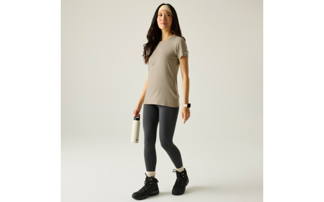 Person in beigem T‑Shirt, schwarzen Leggings und schwarzen Wanderschuhen, hält eine weiße Trinkflasche in einer Hand, trägt Stirnband und Smartwatch, steht auf hellem Hintergrund