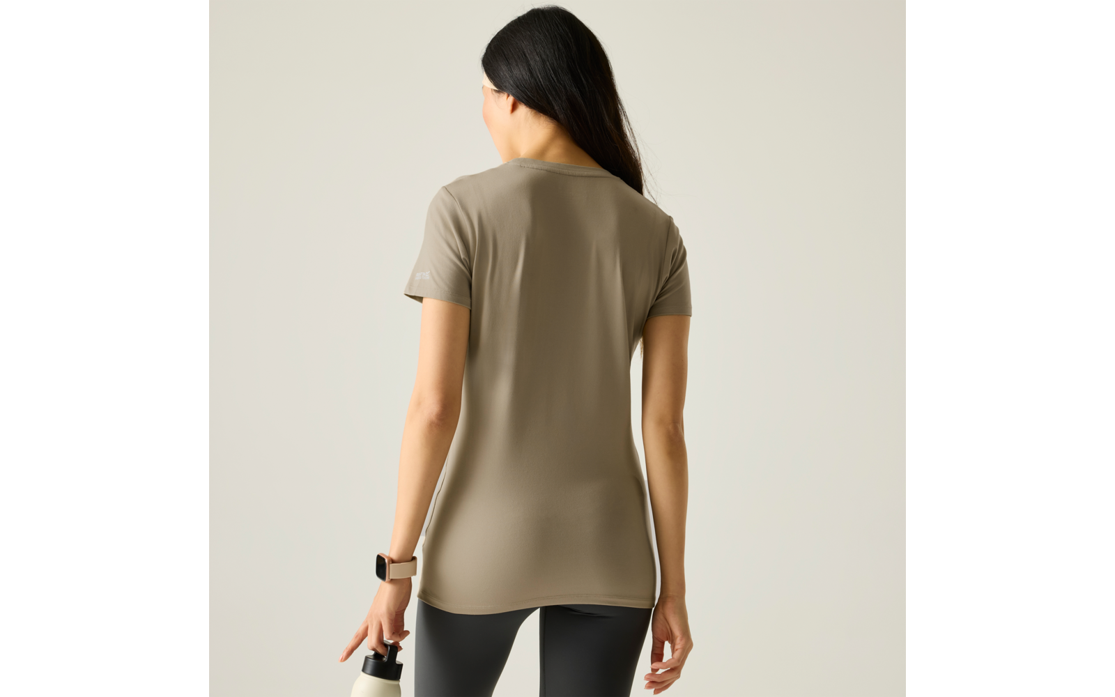 Person von hinten mit langen dunklen Haaren, trägt beige T‑Shirt und schwarze Leggings, hält eine Trinkflasche und trägt eine Armbanduhr vor neutralem Hintergrund