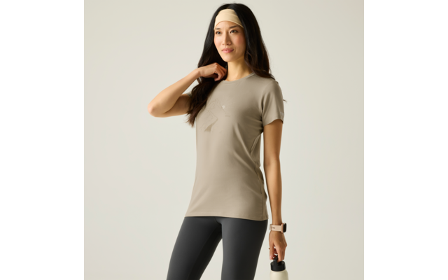 Frau mit langem dunklem Haar und hellem Stirnband, trägt beiges T‑Shirt mit kleinem Aufdruck, dunkle Leggings, hält eine weiße Trinkflasche in der rechten Hand, steht vor hellem Hintergrund