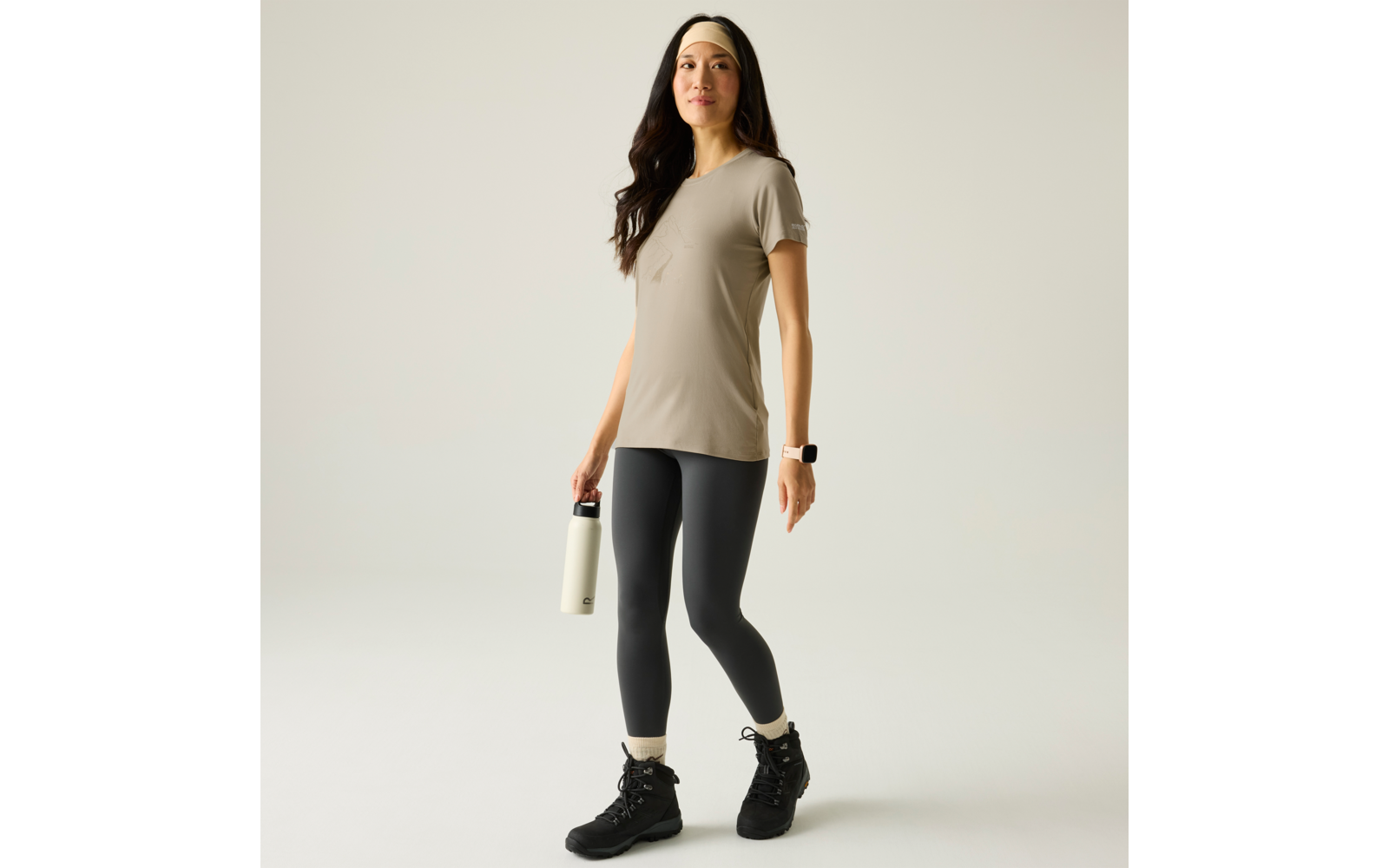 Person in beigem T‑Shirt, schwarzen Leggings und schwarzen Wanderschuhen, hält eine weiße Trinkflasche in einer Hand, trägt Stirnband und Smartwatch, steht auf hellem Hintergrund