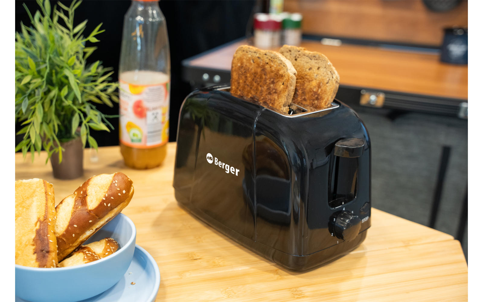 Schwarzer Toaster auf Holzarbeitsplatte mit zwei Toastscheiben im Schlitz; links kleine Topfpflanze, Flasche Saft und Schüssel mit Laugenbrotstücken