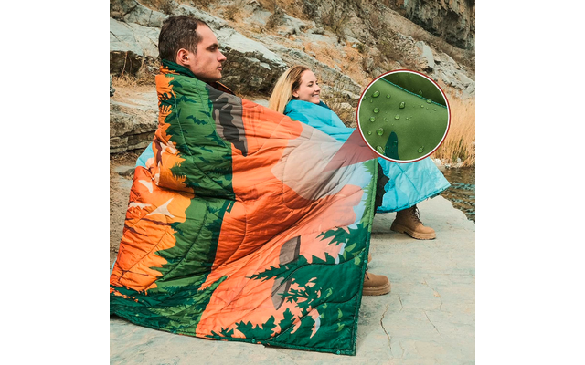 Zwei Personen sitzen auf dem Boden, eingewickelt in eine orange-grüne Decke mit Baum-Motiv, im Freien vor einer Felswand