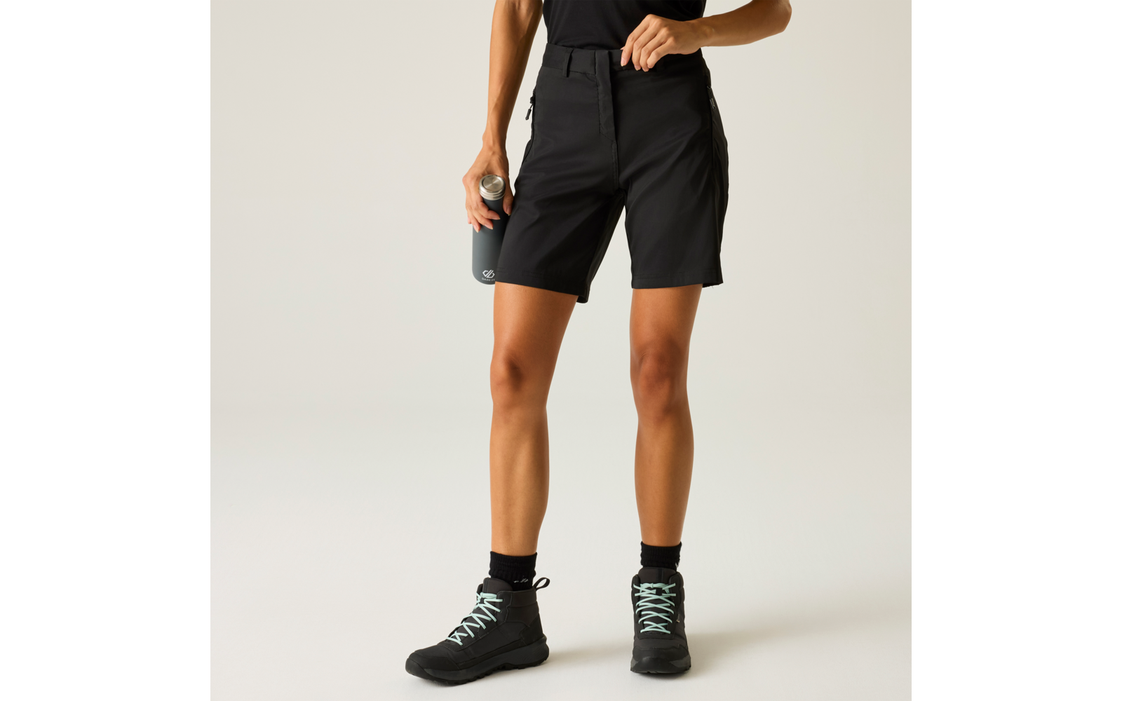 Unterkörper und Beine einer Person in kurzen schwarzen Shorts, schwarze Socken, schwarze Schuhe mit hellen Schnürsenkeln, graue Trinkflasche in der Hand, heller einfarbiger Hintergrund