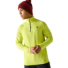 Personne portant un bonnet en tricot rouge à motif avec pompon et un haut manches longues jaune néon à fermeture éclair courte, petits logos sur poitrine et ourlet, mains jointes, regarde vers la gauche, pantalon foncé, fond blanc