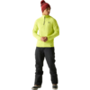 Personne debout, regarde vers la droite, porte une veste jaune fluo à demi-fermeture éclair, pantalon noir, bonnet tricot bordeaux avec pompon et bottes foncées; mains tenues devant le corps