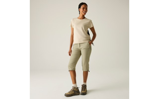 Person steht vor hellem Hintergrund, trägt beige Kurzarmshirt, hellgrüne dreiviertellange Hosen mit Kordeln am Saum, Wanderschuhe und helle Socken; eine Hand in der Hosentasche