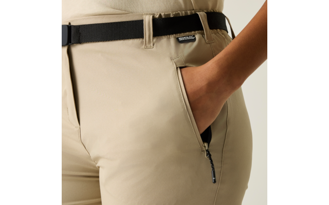 Hüftbereich mit beigen Hosen, rechte Hand in seitlicher Reißverschlusstasche, schwarzer Gürtel und kleines Label am Bund