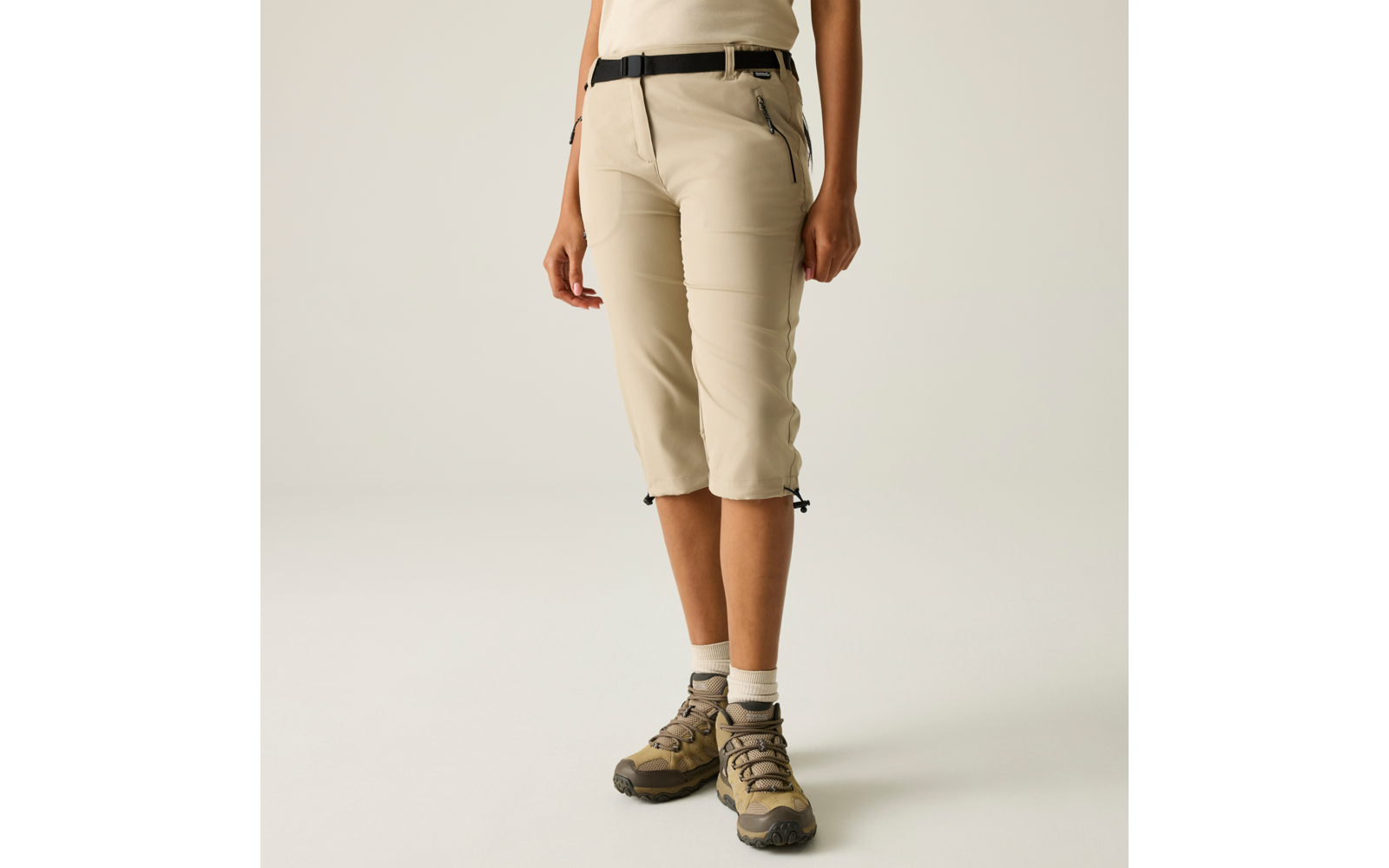 Unterkörper einer Person in knielanger beiger Hose, schwarzem Gürtel, weißen Socken und braunen Schuhen, Hände seitlich, heller Hintergrund