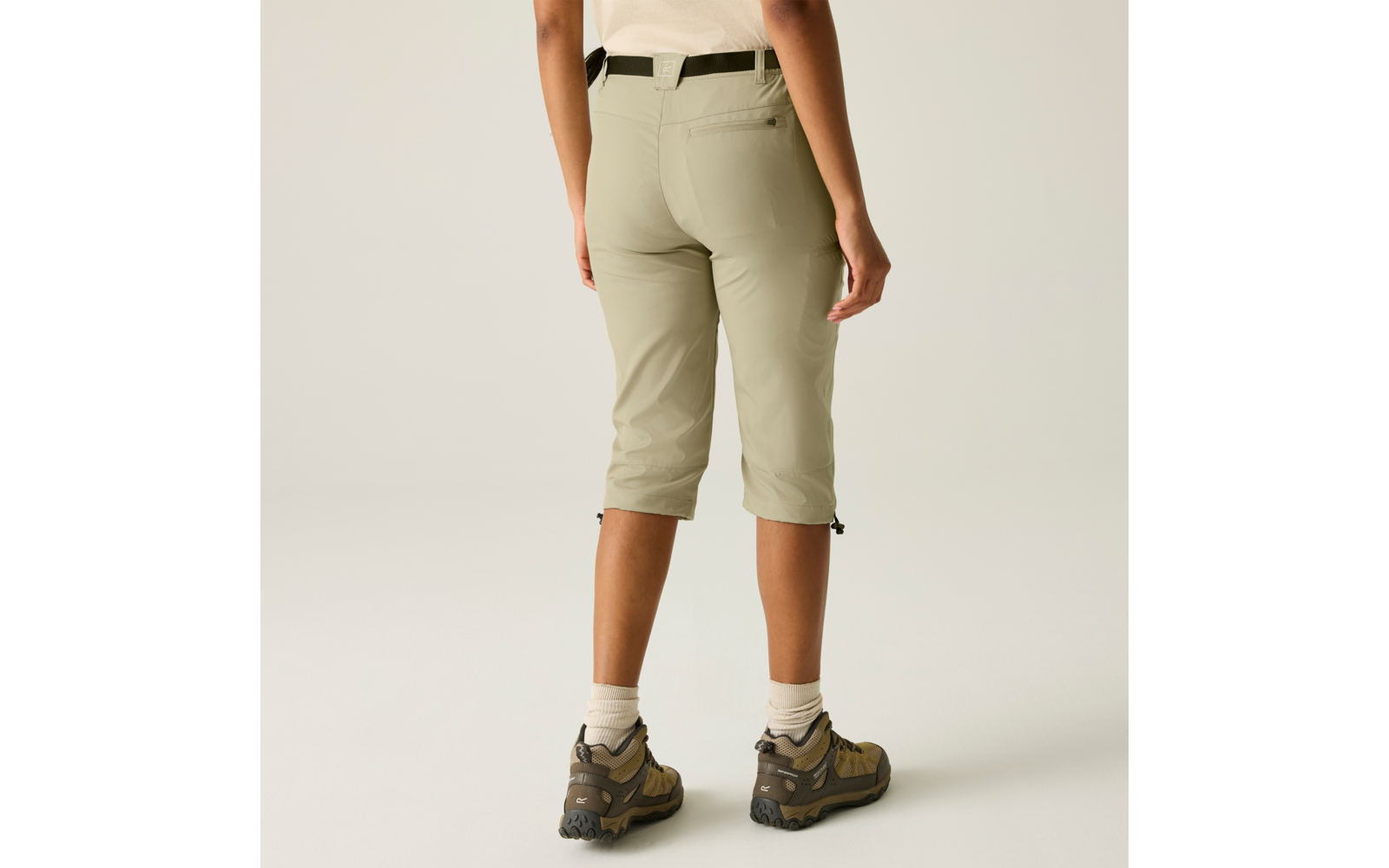 Rückenansicht einer Person von der Taille abwärts, hellgrüne knielange Hose, schwarzer Gürtel, weiße Socken und braune Wanderschuhe vor hellem Hintergrund