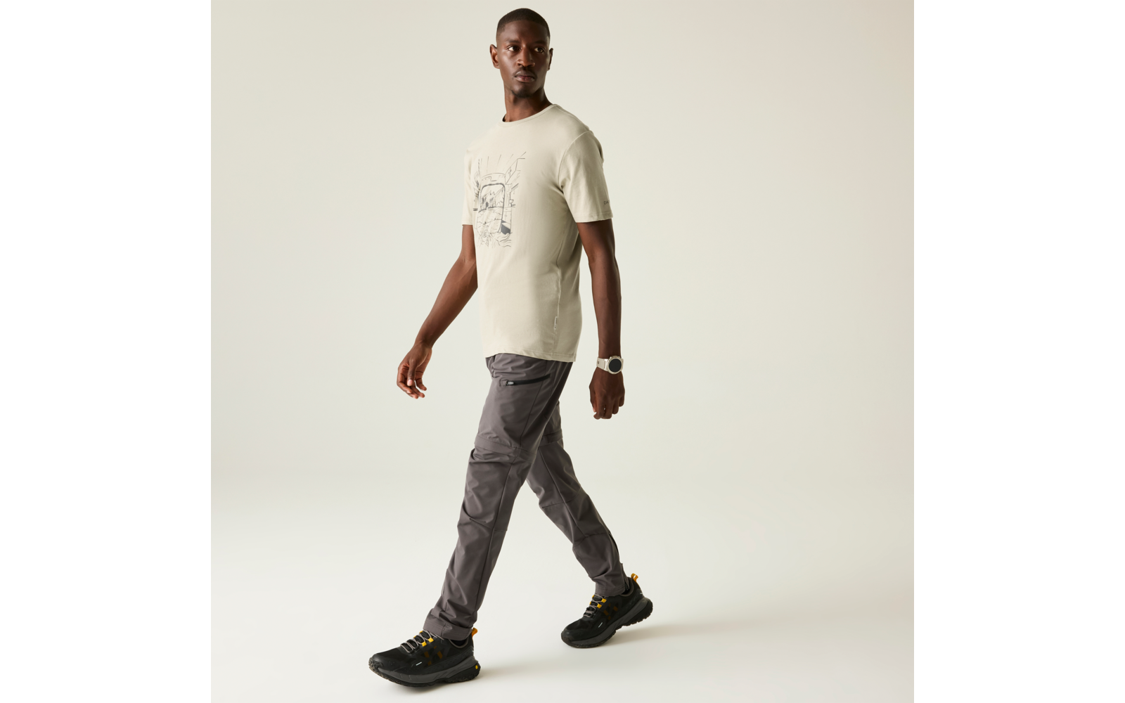 Person im Profil, geht; trägt helles T‑Shirt mit Aufdruck, graue Cargohose und schwarze Sneaker vor hellem Hintergrund