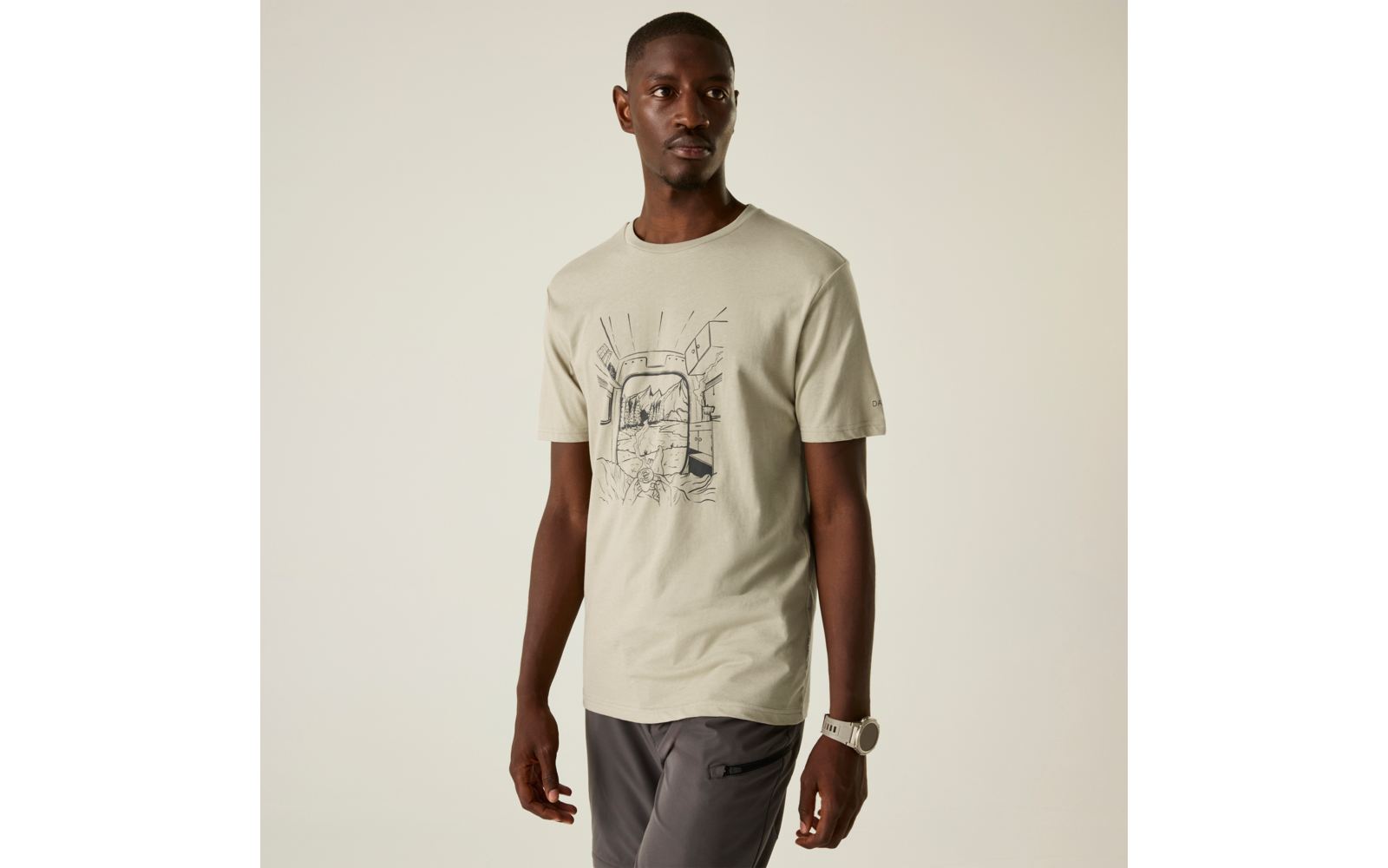 Mann steht vor neutralem Hintergrund, trägt hellbeiges T‑Shirt mit schwarzer Linienzeichnung vorn, dunkelgraue Hose und weiße Armbanduhr am linken Handgelenk