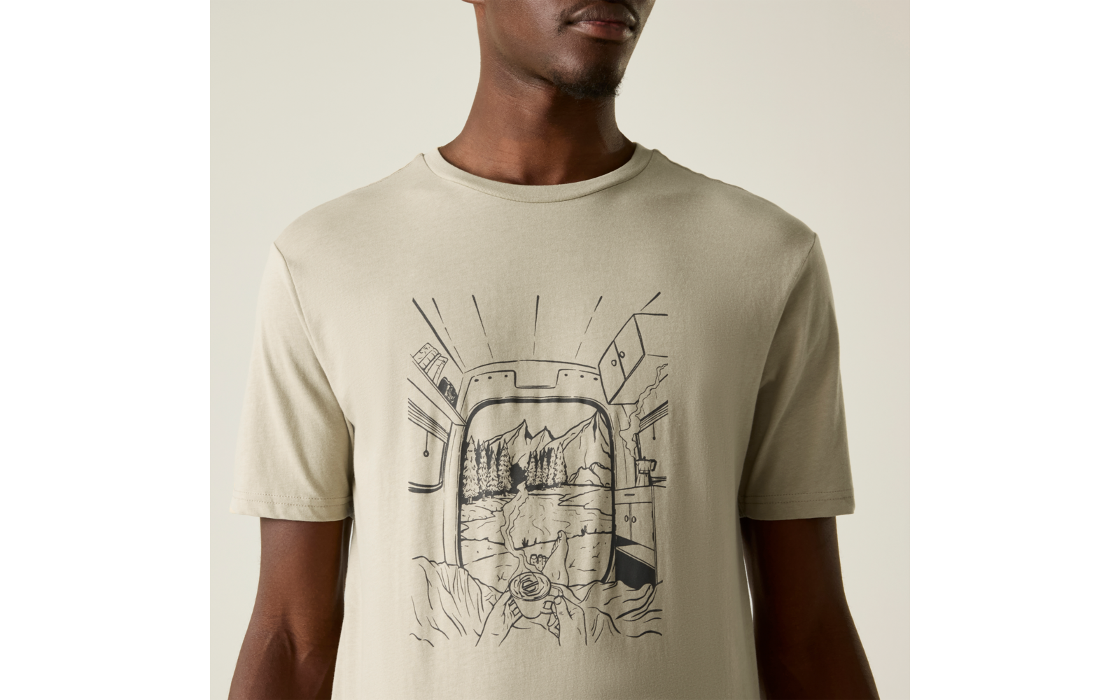 Oberkörper einer Person in beige T‑Shirt mit schwarzer Linienzeichnung: abgerundeter rechteckiger Rahmen, Berglandschaft mit See und Bäumen, felsiger Vordergrund und eine Hand, die eine Kamera hält