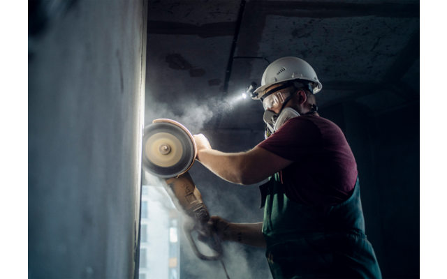 Mann mit Schutzhelm und Maske schleift an einer Wand mit einer Handschleifmaschine