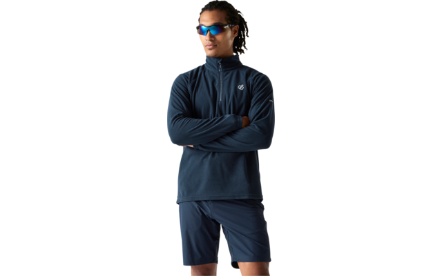 Persoon met donker krullend haar draagt blauwe sportzonnebril, donkerblauw sportjack met korte rits en donkerblau short, staat met gekruiste armen voor een witte achtergrond