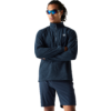 Persoon met donker krullend haar draagt blauwe sportzonnebril, donkerblauw sportjack met korte rits en donkerblau short, staat met gekruiste armen voor een witte achtergrond