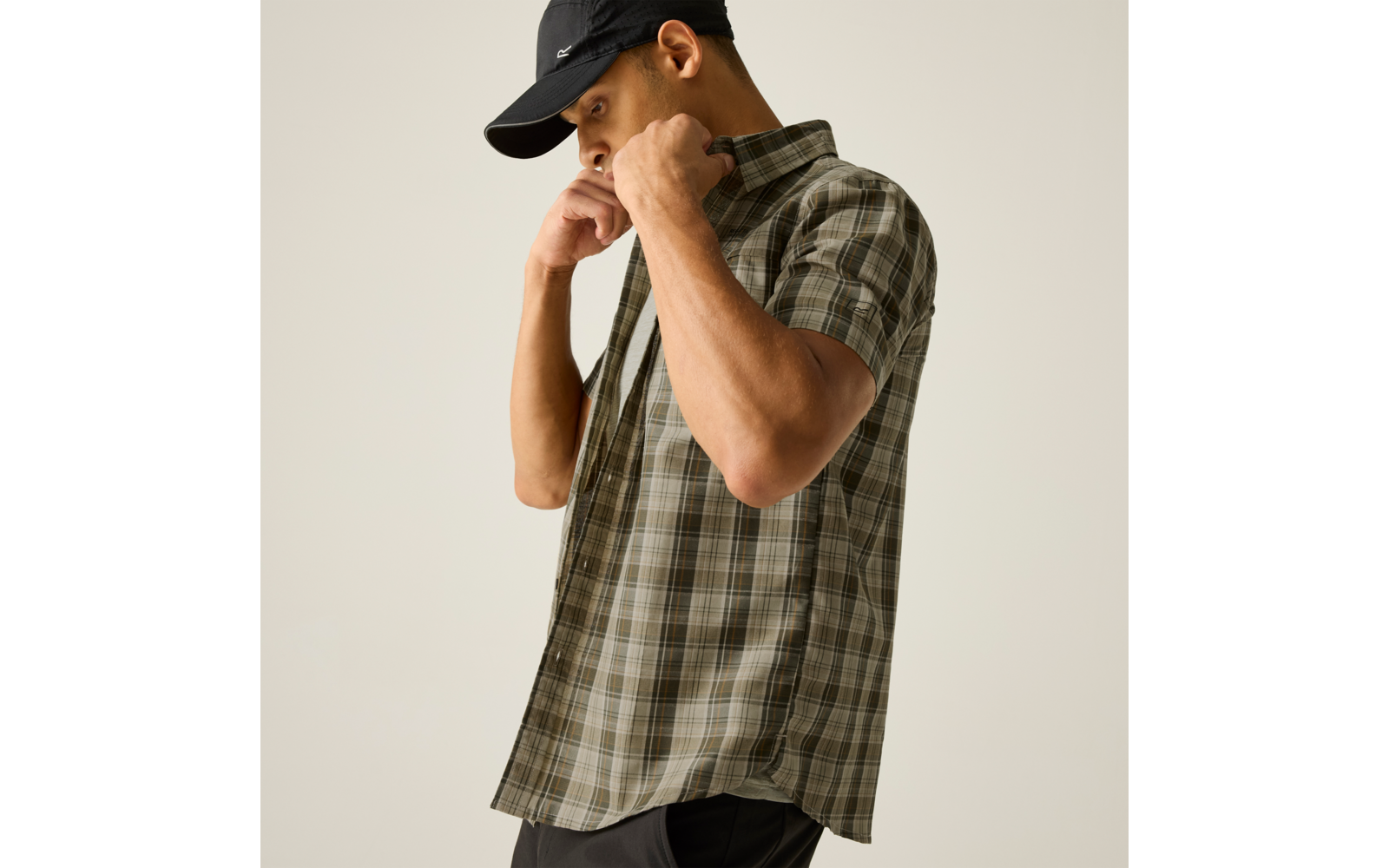 Seitlich stehende Person mit schwarzer Baseballkappe, kurzärmligem kariertem Hemd und dunkler Hose, zieht am Hemdkragen vor hellem Hintergrund