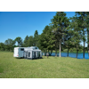 Wohnwagen mit Vorzelt auf einer Wiese neben einem See, zwei Personen stehen davor, Bäume und blauer Himmel im Hintergrund