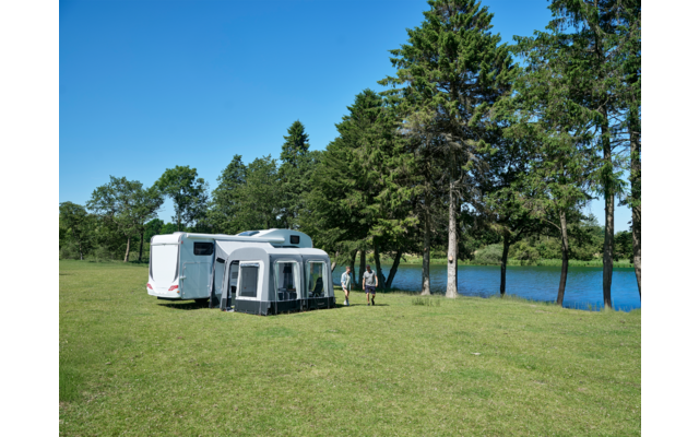 Wohnwagen mit Vorzelt auf einer Wiese neben einem See, zwei Personen stehen davor, Bäume und blauer Himmel im Hintergrund