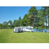 Weißes Wohnmobil mit grauem Vorzelt auf grüner Wiese neben einem See, im Hintergrund Bäume und blauer Himmel
